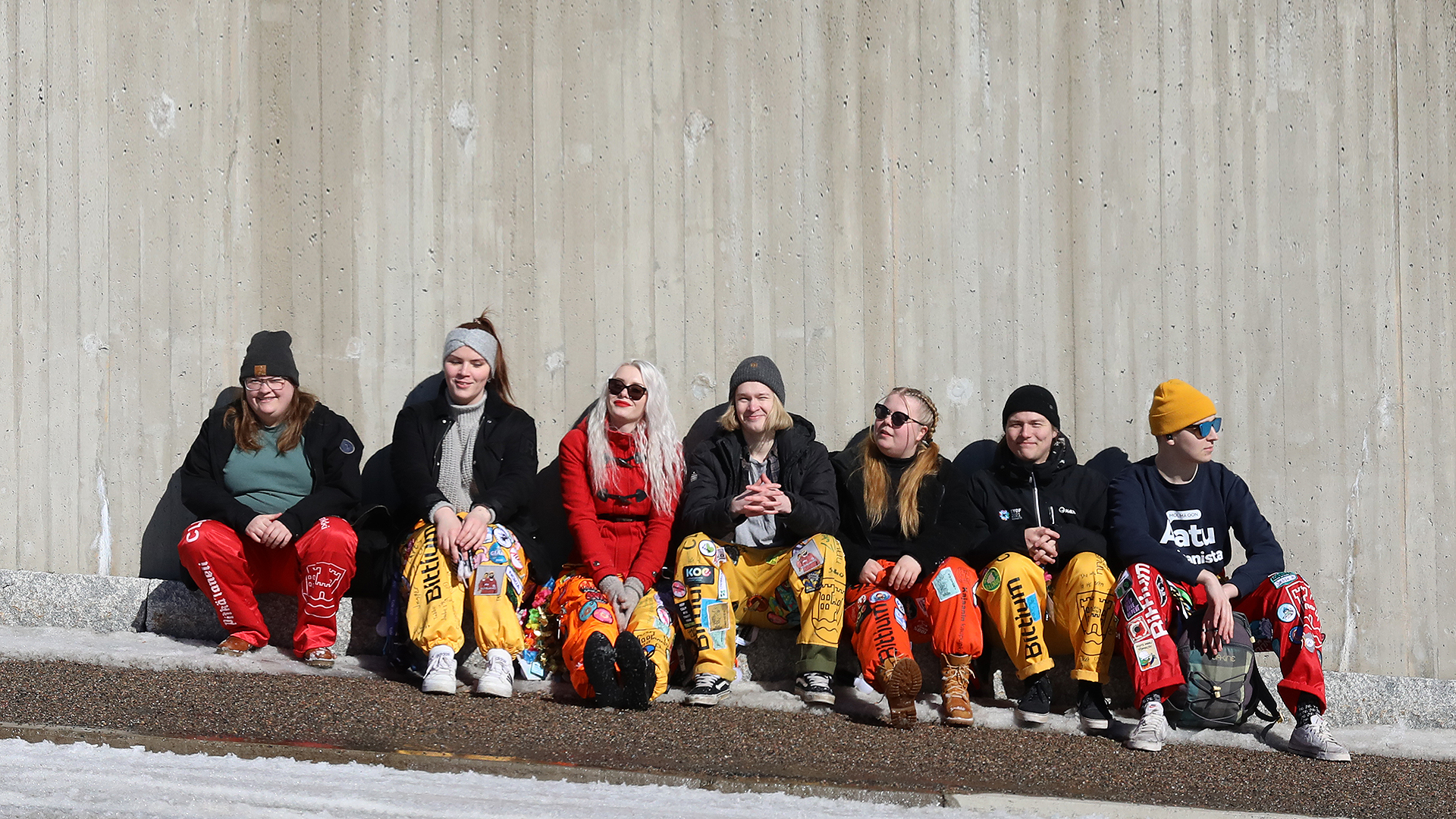 Rivi haalaripukuisia opiskelijoita istuu rivissä tienpenkalla auringossa iloisen näköisenä.
