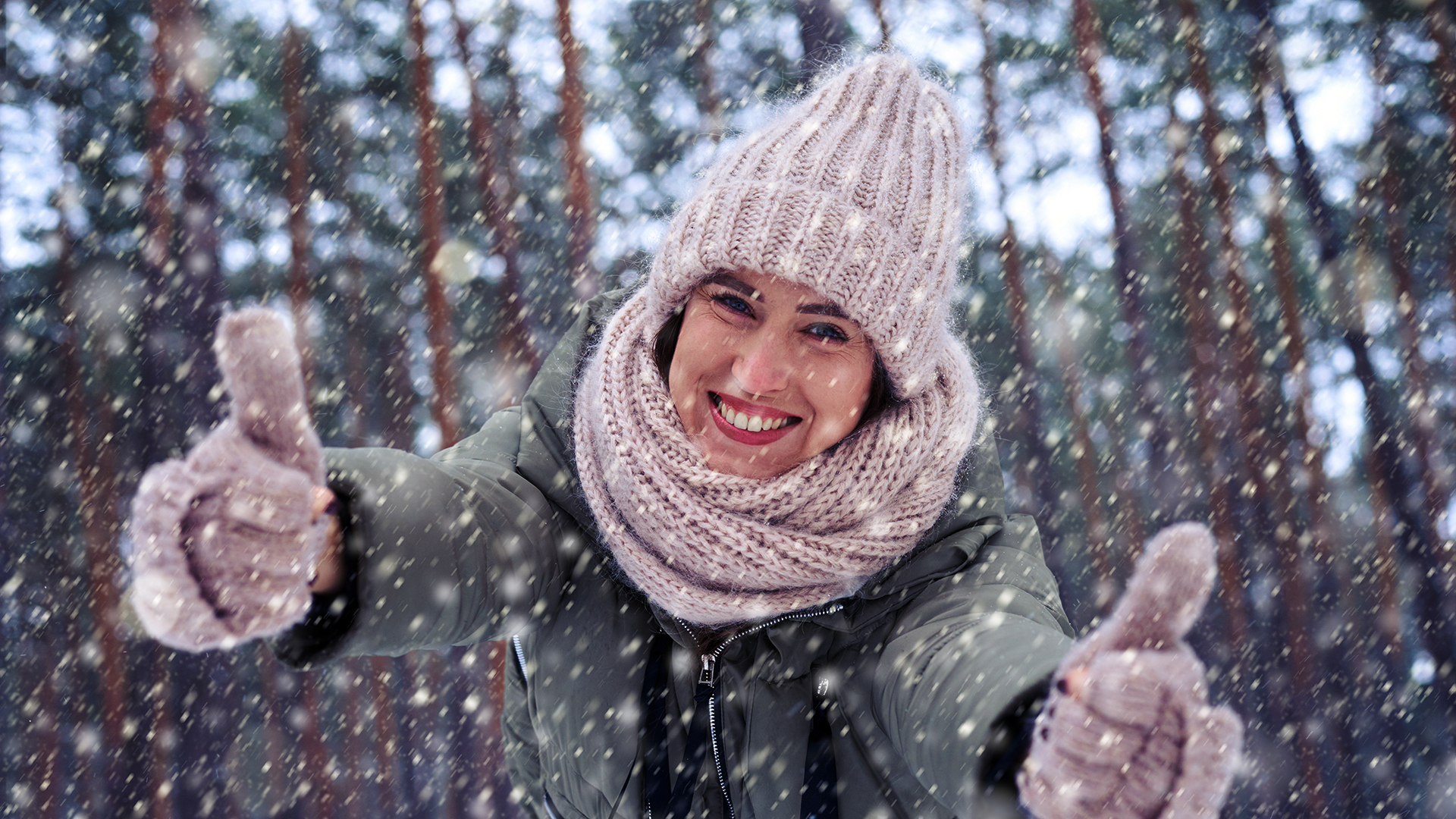 Pipopäinen nainen seisoo lumisateessa ja hymyilee, sekä näyttää peukkua.